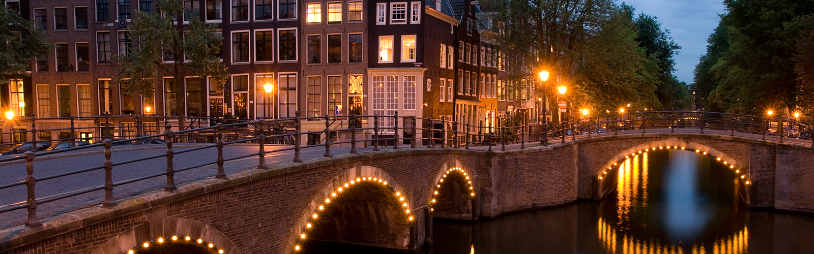 A bridge covered in lights over a river in front of several narrow buildings, glowing in the twilight. 